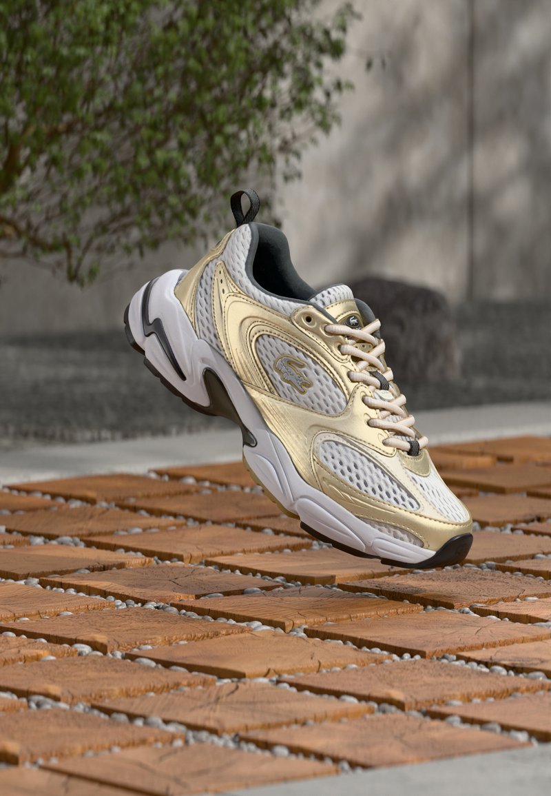 Λευκό και μπεζ παπούτσι με λεπτομέρειες από δίχτυ και δέρμα, αιωρούμενο πάνω από καφέ ξύλινη επιφάνεια με θολό πράσινο φυτό και γκρι τοίχο στο φόντο.