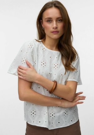 Femme aux longs cheveux bruns portant un chemisier blanc brodé à manches courtes et un bracelet marron, bras croisés devant un fond uni.