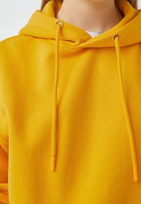Gros plan d'une personne portant un sweat à capuche jaune vif avec des cordons et un col de chemise blanc visible au niveau du cou.