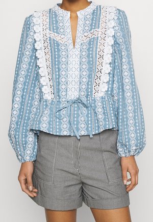 Blouse bleue à manches longues avec motif, détails en dentelle blanche et ceinture à nouer, associée à un short rayé vertical noir et blanc.