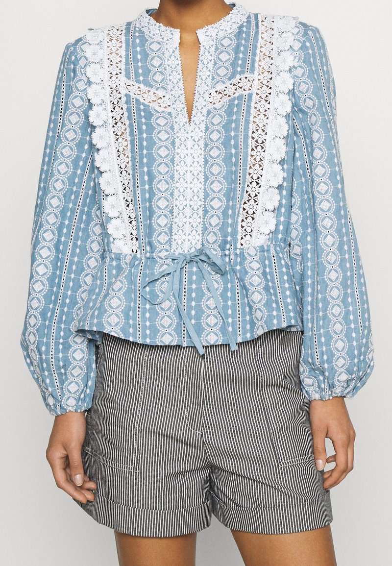 Blouse bleue à manches longues avec motif, détails en dentelle blanche et ceinture à nouer, associée à un short rayé vertical noir et blanc.