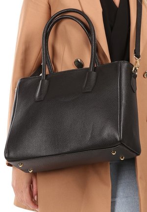 Sac à main en cuir noir avec deux poignées supérieures, de forme rectangulaire, surface texturée et accents en métal doré, reposant contre un manteau beige.