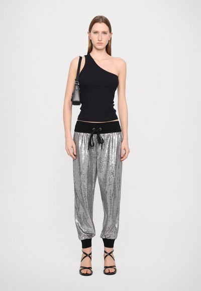 Zwarte een-schouder fitted top gecombineerd met glanzende zilveren joggerpants met elastische boorden en een zwarte tailleband. Model draagt gespen met zwarte hakken.