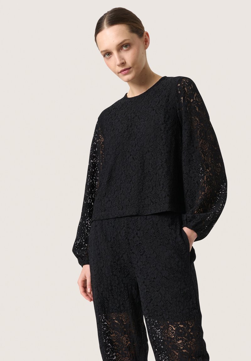 Top in pizzo nero con maniche lunghe e un fit rilassato, caratterizzato da un motivo floreale testurizzato. Abbinato a pantaloni in pizzo coordinati, entrambi con tasche.