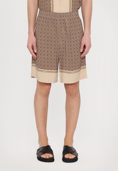 Braun gemusterte Shorts mit einem elastischen Bund, versehen mit einem beigen Saum. Tragen zu schwarzen Schlappen.