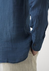Hombre vistiendo una camisa de manga larga azul marino con puños abotonados y pantalones beige, con una mano en el bolsillo.