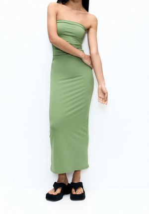 Femme portant une robe longue bustier vert olive avec un détail froncé, associée à des sandales à plateforme noires, debout sur un fond blanc.