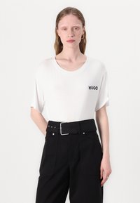 Witte katoenen t-shirt met een ronde halslijn en korte mouwen, met het woord "HUGO" in het zwart op de linkerborst. Draagt het met zwarte broek met riem.
