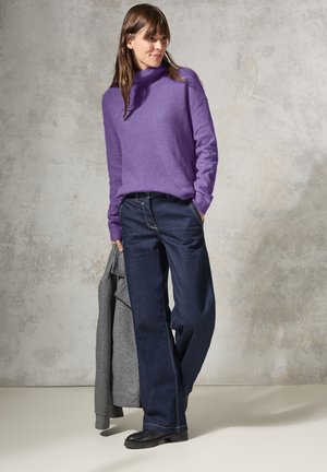 Purple knit sweater with a high neck, paired with dark blue wide-leg jeans. A grey jacket draped over one arm, black shoes complete the outfit.