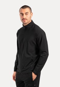 Schwarze ZIP-Jacke aus glattem, leichtem Stoff; mit hohem Kragen und Seitentaschen. Getragen mit schwarzen Hosen.