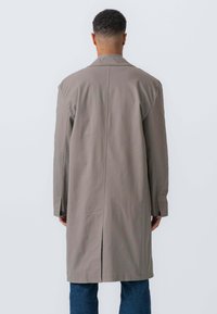 Lang, lysegrå frakke med slids bagpå og knaplukning ved ærmerne, lavet af glat stof. Båret over blå jeans, set fra ryggen.