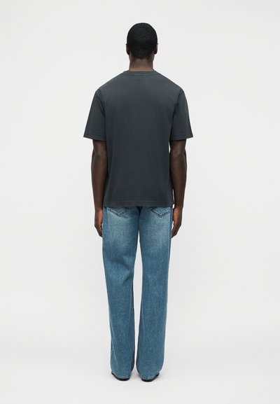 Άνδρας που στέκεται με την πλάτη γυρισμένη, φορώντας ένα μονόχρωμο σκούρο γκρι κοντομάνικο μπλουζάκι και άνετο ανοιχτό μπλε τζιν, μπροστά σε λευκό φόντο.