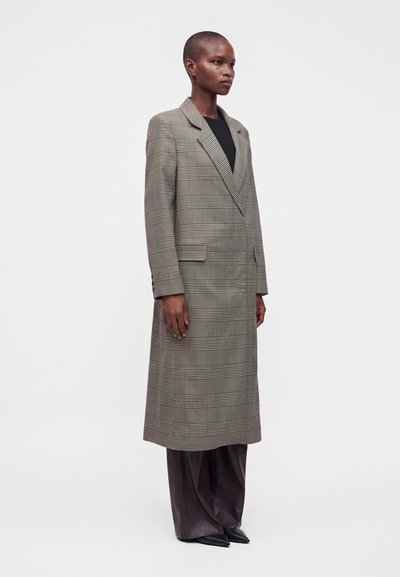 Personne grande portant un long manteau à carreaux par-dessus un haut noir et un pantalon ample foncé, debout devant un fond blanc uni.