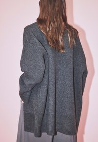 Osoba s dlhými hnedými vlasmi, oblečená v prehnane veľkom tmavosivom pletenom svetri a sivých nohaviciach, stojí pred svetloružovým pozadím.