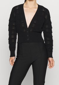 Svart stickad cardigan med djup V-ringning, vågmönster med öppna maskor, ribbad fåll och fyra svarta knappar framtill.
