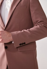 Hellrosa Blazer mit schlankem Design, einer einzelnen Brusttasche und drei schwarzen Knöpfen am Ärmel. Glatte Stofftextur und taillierte Passform.