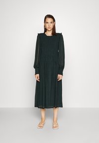 Vestido verde de mangas compridas com corpete franzido e ombros rufados, apresentando uma saia fluida e um padrão subtil. Usado com sandálias brancas de tiras.