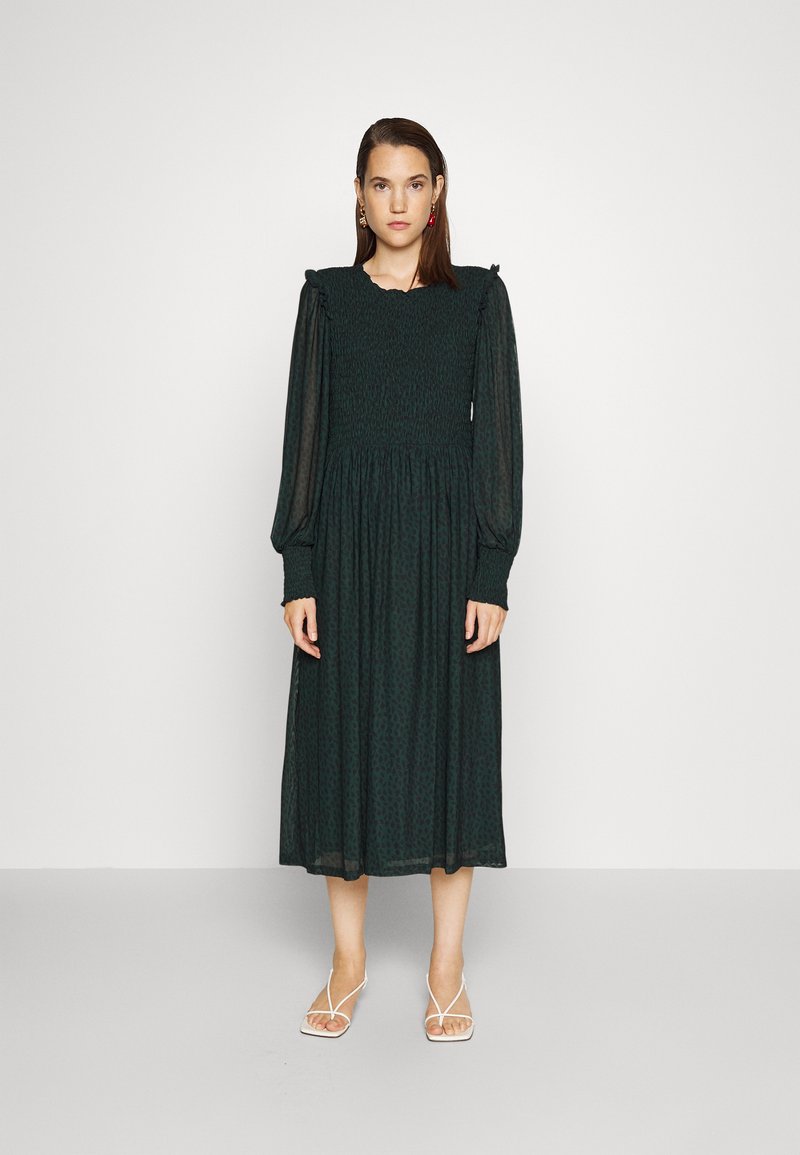 Vestido verde de mangas compridas com corpete franzido e ombros rufados, apresentando uma saia fluida e um padrão subtil. Usado com sandálias brancas de tiras.