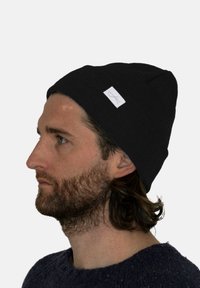 Black knit beanie featuring a turned-up cuff and a small white tag displaying a logo, worn on a model with long hair and a beard.