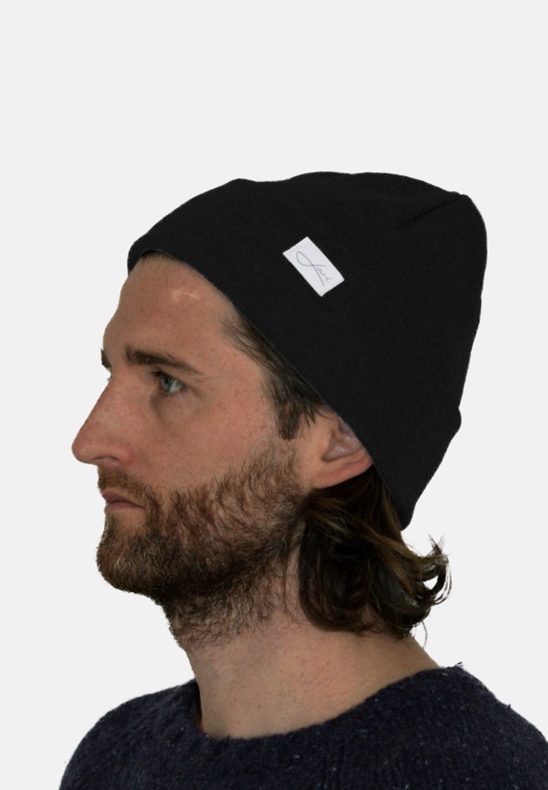 Black knit beanie featuring a turned-up cuff and a small white tag displaying a logo, worn on a model with long hair and a beard.