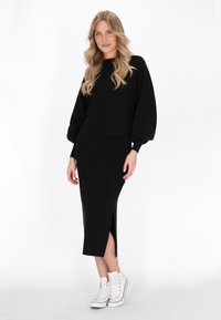 Černý pletený crop top s nafouknutými rukávy v kombinaci s černou midi sukní s bočním rozparkem, doplněný bílými teniskami na vysoké podrážce.