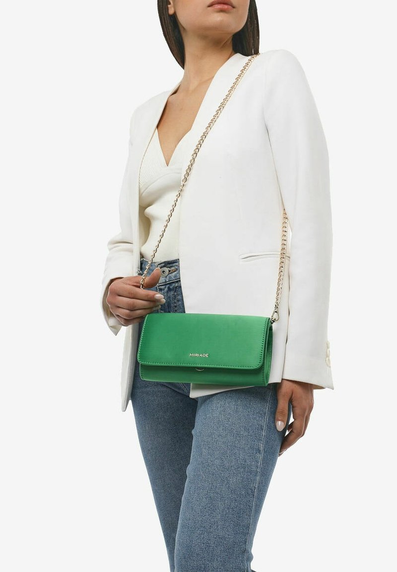 Borsa a tracolla verde in pelle con una catena dorata, dotata di chiusura a pattina e dettaglio con logo, abbinata a un blazer bianco e jeans.