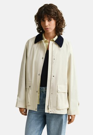 Vrouw met krullend haar, gekleed in een crèmekleurig jack met een zwarte kraag, een geel gestreept overhemd, een zwart shirt en een blauwe spijkerbroek tegen een effen achtergrond.