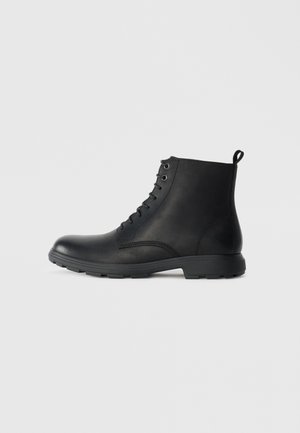 Botas de cuero negro hasta el tobillo con puntera redondeada, diseño de cordones y acentos texturizados. Suela de goma con perfil bajo y tira en la parte trasera.