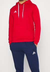 Roter Hoodie mit weißen Kordeln, Kängurutasche, Adidas-Logo. Kombiniert mit dunkelblauen Jogginghosen mit weißen Logoakzenten.