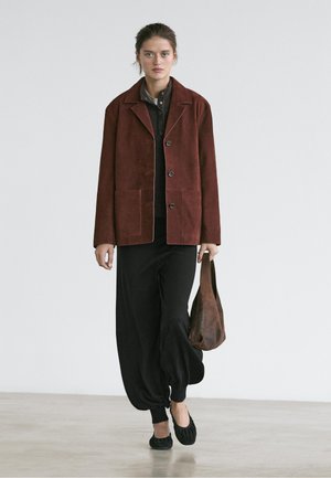 Kobieta ubrana w rdzawy zamszowy płaszcz, czarne luźne spodnie, czarne baleriny, niosąca brązową skórzaną torebkę, stojąca na jasnej podłodze na białym tle.