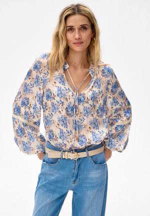 Vrouw met golvend blond haar, gekleed in een losse crèmekleurige blouse met blauwe bloemenprint, lichte beige riem en lichtblauwe spijkerbroek met hoge taille tegen een witte achtergrond.