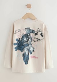 Camicia beige a maniche lunghe con una grande grafica di un robot blu e grigio, con contorni neri. Testo "Transformers" in rosa nella parte inferiore.