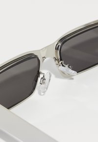 Sonnenbrille mit auffälligen, rechteckigen schwarzen Gläsern, einem silbernen Metallrahmen und klaren Silikonnasenpads. Mit Scharnierdetails und einem eleganten Design.