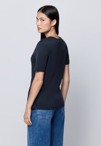 Giovane donna con capelli lunghi e scuri, che indossa una maglietta nera a maniche corte e jeans di denim blu, in piedi contro uno sfondo chiaro e semplice.