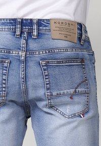 Los jeans de mezclilla azul claro presentan una etiqueta de cuero marrón con logo, costuras en contraste y dos bolsillos traseros con un acento rojo.