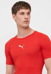 Rød treningsskjorte med korte ermer, laget av glatt stoff. Har en hvit Puma-logo på brystet. Designet for aktiv bruk.