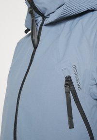 Chaqueta de color azul claro con cremalleras y acentos en negro. Cuenta con una capucha texturizada y un logotipo de la marca bordado en el frente. Acabado de tela suave.