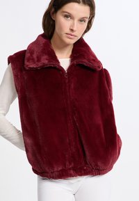 Gilet in ecopelliccia borgogna con zip frontale, colletto e vita elasticizzata. Presenta un design senza maniche e una texture morbida.