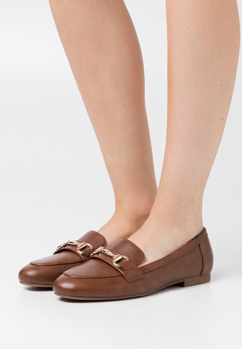 Mocassins en cuir brun avec une semelle plate, présentant un accent de chaîne dorée à l'avant et une finition lisse.
