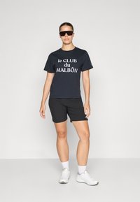 T-shirt in cotone nero con scritta bianca "le CLUB du MALBÔN", abbinato a pantaloni corti neri e sneakers bianche chunky, indossato con occhiali da sole scuri.