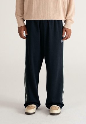 Person wearing black wide-leg track pants with white side stripes, beige knitted sweater, and beige shoes with woven top detail.