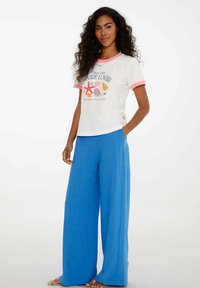 Žena s dlouhými vlnitými vlasy, oblečená v bílém tričku s potiskem mušlí a modrých širokých kalhotách, stojí s jednou rukou v kapse.