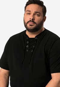 Dospělý muž s vousy, oblečený v černé košili s krátkým rukávem a šněrovacími detaily, hledí mírně na stranu na jednolitém pozadí.