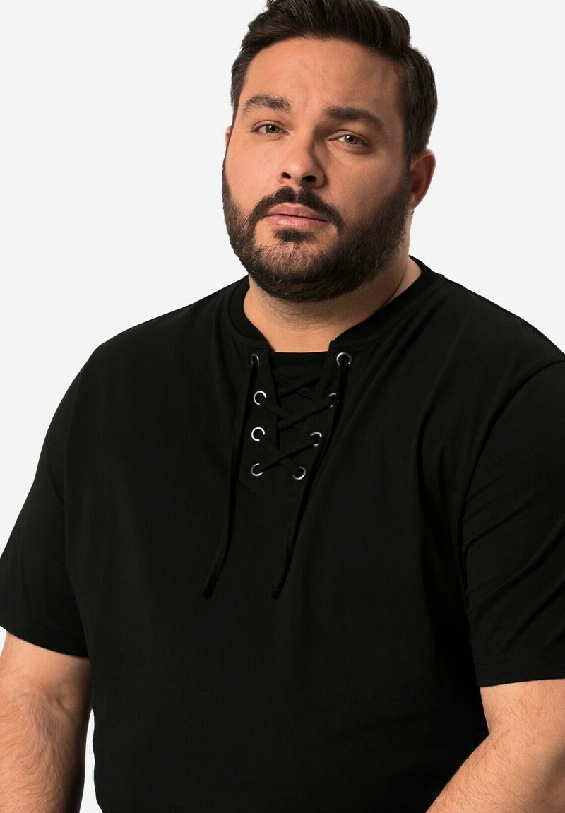 Dospělý muž s vousy, oblečený v černé košili s krátkým rukávem a šněrovacími detaily, hledí mírně na stranu na jednolitém pozadí.