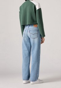 Persona con jeans rectos de cintura alta en azul claro, sudadera corta verde con paneles blancos en los hombros y zapatillas blancas, de pie de espaldas.
