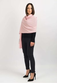 Lyserød sjal draperet over en skulder, lavet af blødt stof. Båret over et mørkt outfit med tætsiddende sorte bukser og højhælede sko.