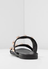 Black flat sandals with two straps, featuring metallic rose gold studs. The straps are made of smooth leather, with a flat rubber sole.
