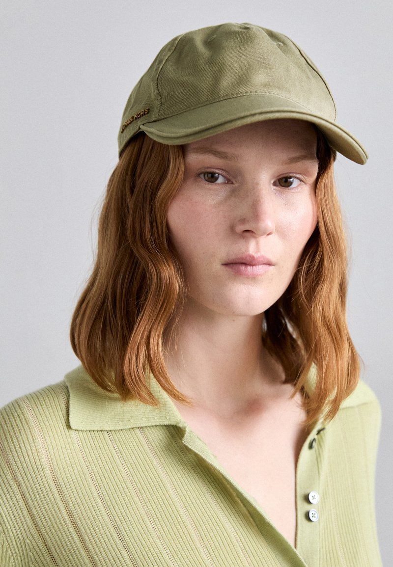 Cappello verde oliva con visiera pre-curvata e tessuto texture. Presenta un dettaglio in metallo dorato. Il modello indossa un top a coste verde chiaro con bottoni.