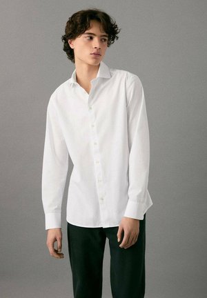 Giovane con capelli ricci scuri che indossa una camicia bianca con bottoni e pantaloni neri, in piedi davanti a uno sfondo grigio uniforme.