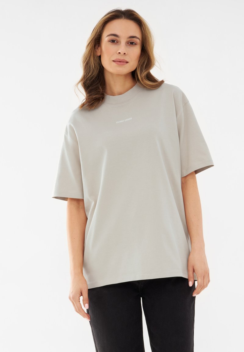 Világosszürke, túlszíjazott póló, sima pamutból készült, kerek nyakkivágással és rövid ujjal. Középen felirat található. Fekete nadrág látható.